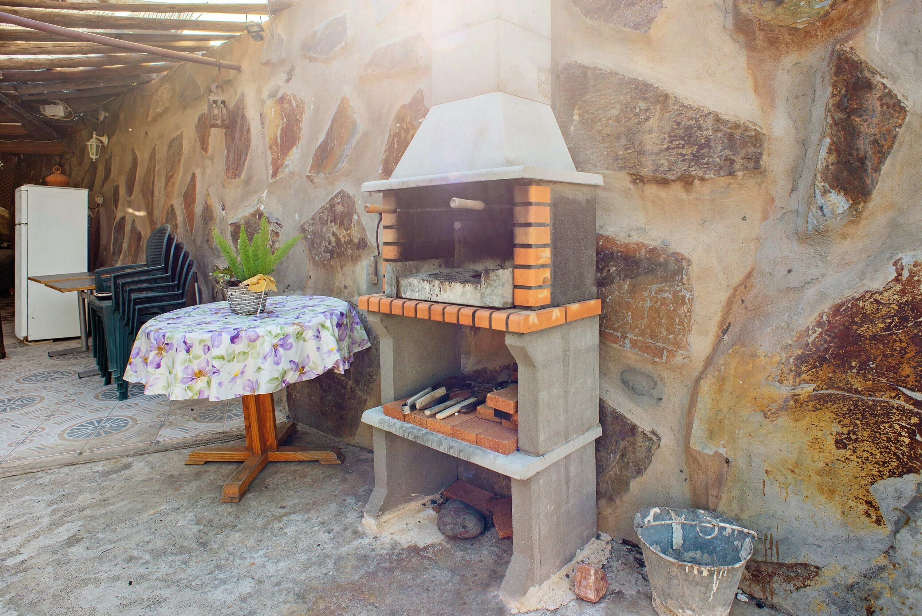 Een stenen muur, een bakoven, een tafel met een bloemrijk tafelkleed en een stoel.