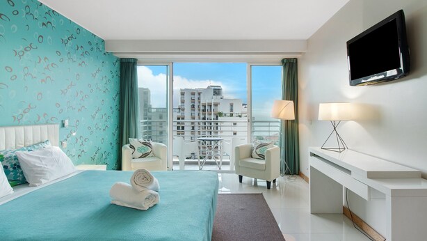 Een hotelkamer met een bed, een bureau, een televisie en een balkon met uitzicht op de stad.