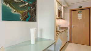 Hotelkamer met glazen tafel, witte vaas, schilderij, kitchenette en houten deur met een nooduitgangsbord.