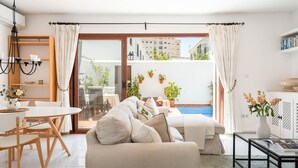 Een moderne woonkamer met een bank, een eettafel en stoelen, een glazen deur die leidt naar een balkon met planten en een zwembad.