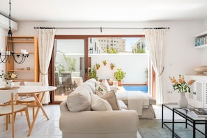 A modern living room with a sofa, dining table, and chairs, a glass door leading to a balcony with plants and a pool.