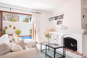 A living room with a sofa, a glass coffee table, a fireplace, a bookshelf, and a view of a building outside.