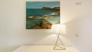 Een schilderij van een kustlandschap aan een muur, een witte tafel met een lamp, en een witte kast eronder.