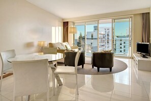 A modern hotel room with a dining area, a sofa set, a TV, and a balcony with a city view.