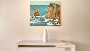 Een schilderij van kliffen aan de kust aan een muur, een wit bureau met een cilindervormig voorwerp erop, en een effen muur.