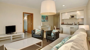 Een moderne woonkamer met een flatscreen televisie, een witte salontafel, twee leren fauteuils, een bank en een keukenhoek met een magnetron.