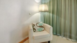 Ein kleiner, weißer Sessel mit einem blumigen Kissen, platziert in der Nähe einer Stehlampe und eines grünen Vorhangs.