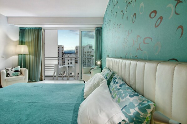 A hotel room with a bed, a chair, a table, and a balcony with a city view.