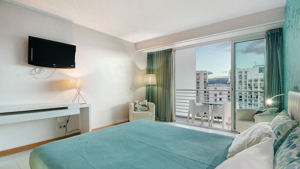 Ein Hotelzimmer mit Bett, Fernseher, Schreibtisch mit Lampe, Stuhl und Balkon mit Blick auf Gebäude.