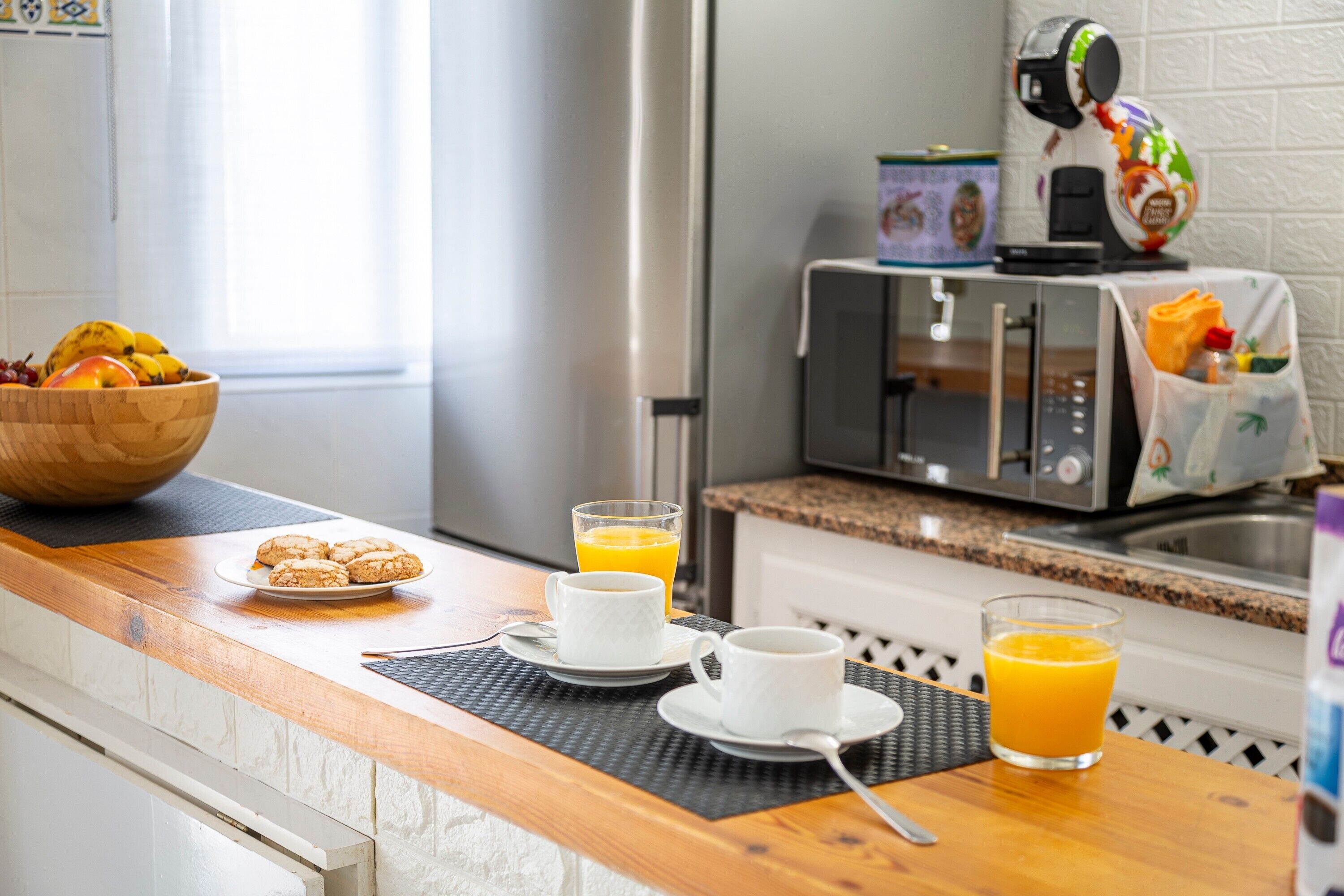 A kitchen counter with a wooden table, a bowl of fruit, two cups of orange juice, and a coffee maker.