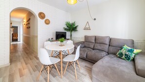 A modern living room with a white brick wall, a brown leather sofa, a round dining table with white chairs, and a hanging light fixture.