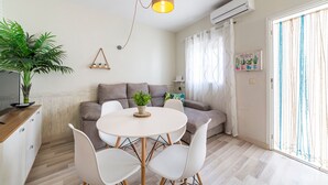 A modern living room with a round dining table, white chairs, a grey sofa, a hanging light fixture, a potted plant, and a window with curtains.