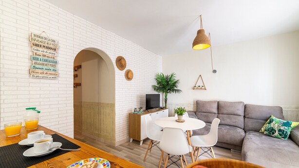 A modern living room with a white brick wall, a grey sofa, a dining area with a table and chairs, and a TV on a wooden cabinet.