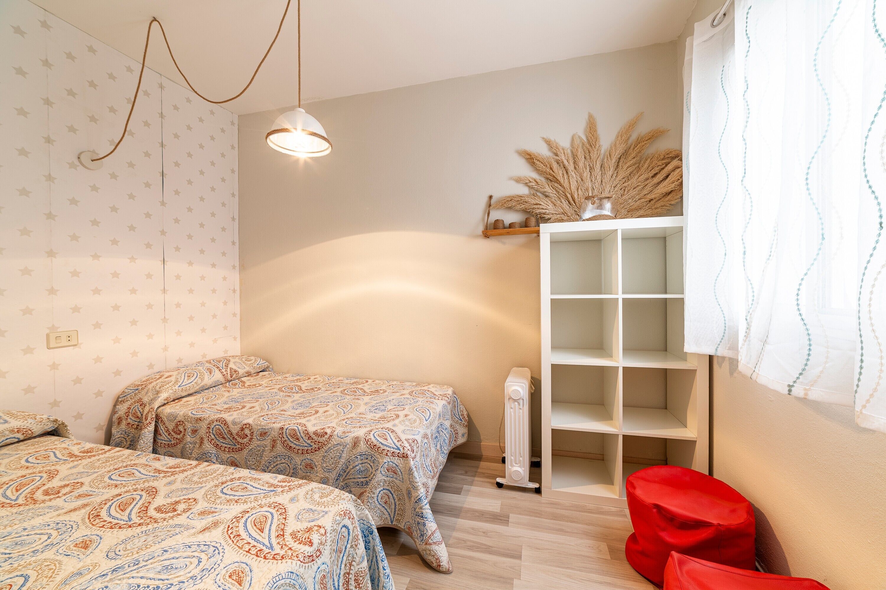 Un dormitorio con dos camas, un radiador, una estantería con plantas decorativas y un cojín rojo.