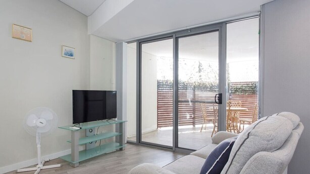 Una sala moderna con televisor de pantalla plana, una mesa de centro de vidrio, un sofá y un ventanal corredero que da a un espacio exterior con mesa y sillas.