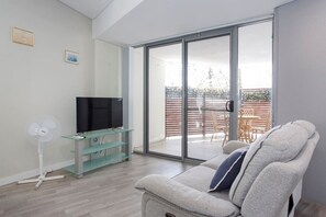 A modern living room with a flat-screen TV, a glass coffee table, a sofa, a sliding glass door leading to an outdoor area with a table and chairs.