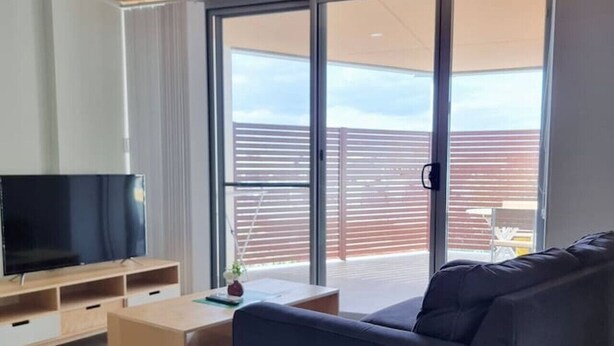 A modern living room with a black sofa, a wooden coffee table, and a TV on a wooden stand. There is a sliding glass door leading to an outdoor area. - Waverley apartment D-one bedroom, wifi available (Beckenham)