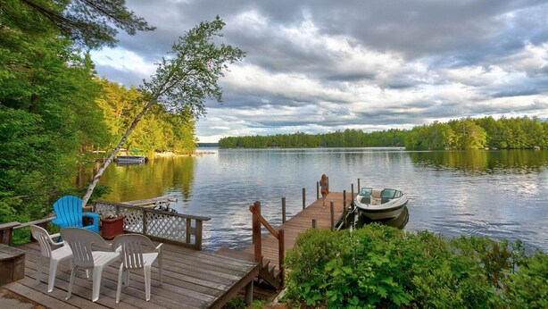 A wooden dock with chairs and a boat, overlooking a calm lake with trees and a cloudy sky. - Winnipesaukee Waterfront Home Sleeping for 8 (Moultonborough)