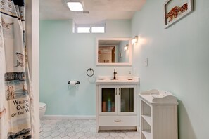 Un baño con un lavamanos blanco, un espejo y un inodoro. Hay una cortina de ducha con una escena de playa. Las paredes están pintadas de azul claro, y el piso tiene baldosas hexagonales.