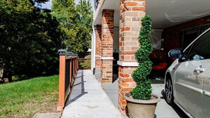 A residential driveway with a white car parked on the right, a brick pillar with a potted plant, and a garage door visible in the background.