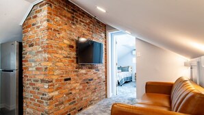 A modern loft bedroom with exposed brick walls, a flat-screen TV mounted on the wall, a built-in wardrobe, and a large window offering a view of the outdoors.