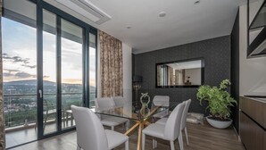 A modern living room with a glass dining table, white chairs, a large potted plant, and a view of a cityscape through large windows. - Africa's Touch & Leisure (Kigali)