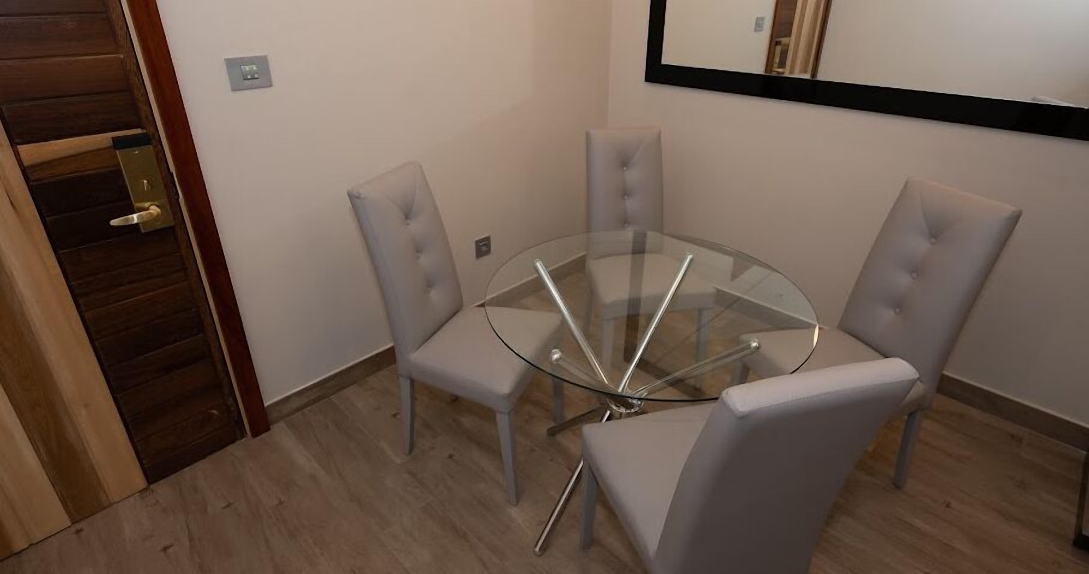 A small, modern dining area with a glass table surrounded by four grey upholstered chairs.