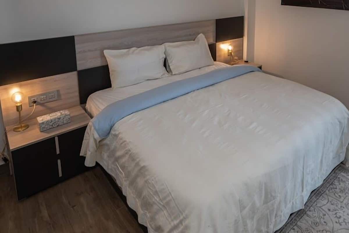 Ein ordentlich bezogenes Bett mit blauem Akzent, ein Nachttisch mit Lampe und eine Wandleuchte.