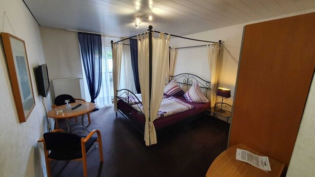 A four-poster bed with a canopy, a small table with a chair, a television on the wall, and a window with curtains. - Hotel Jungmann (Saarlouis)