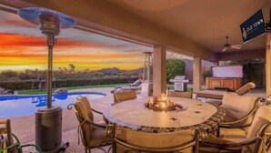 Eine Terrasse mit rundem Tisch und Stühlen, einer Feuerstelle und Blick auf einen Pool und die Landschaft bei Sonnenuntergang.