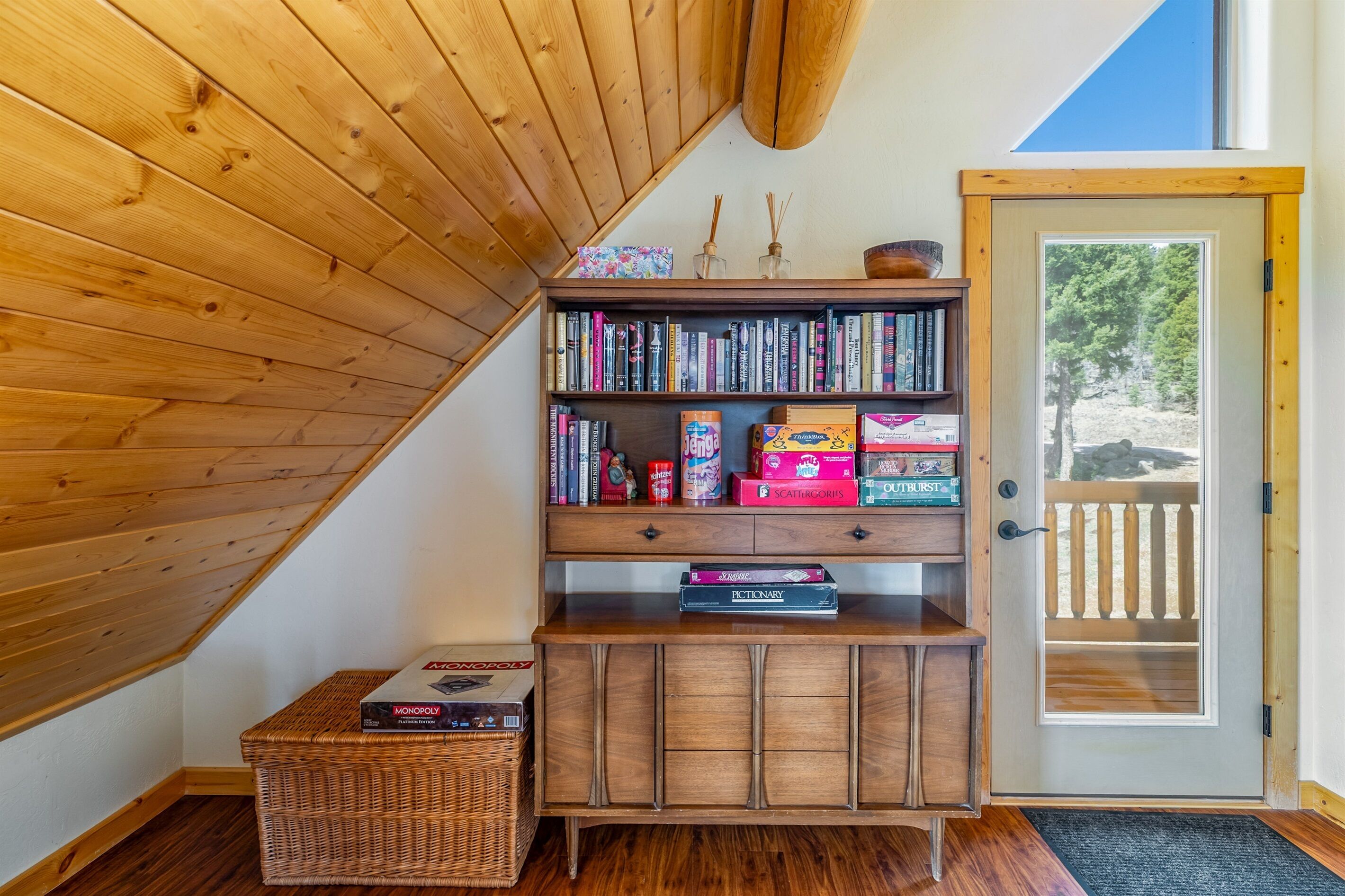 A wooden bookshelf with books and board games, a wicker basket, and a door with a view of trees.