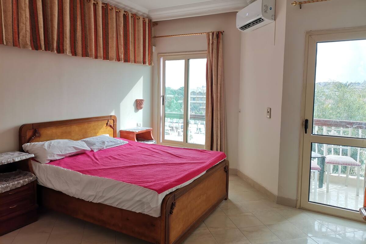 A bedroom with a wooden bed, a nightstand, a small table, a window with curtains, and a view of the outside balcony.
