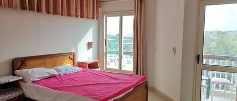 A bedroom with a wooden bed, a nightstand, a small table, a window with curtains, and a view of the outside balcony.
