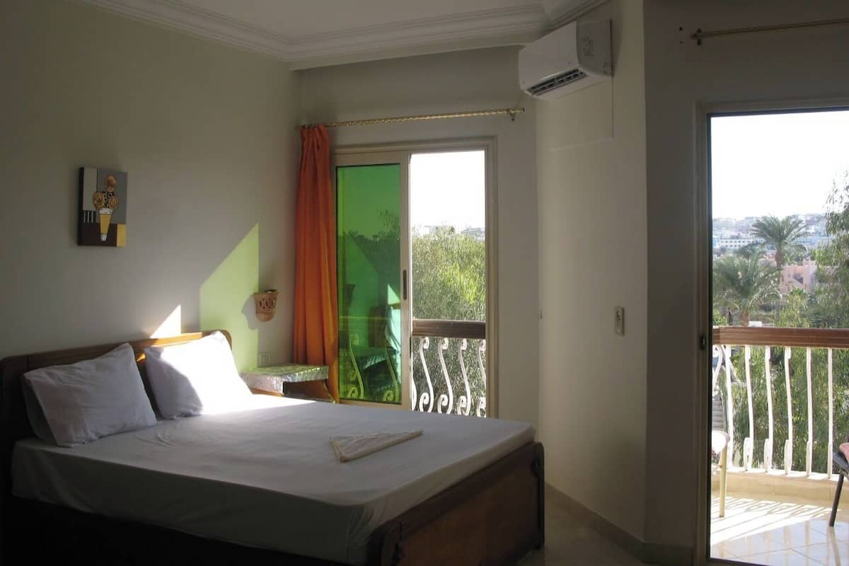A bedroom with a bed, a window, a balcony, and an air conditioning unit.