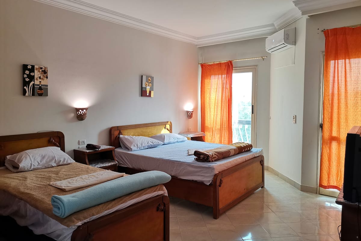 A hotel room with two wooden beds, a nightstand, a wall-mounted air conditioner, and a window with orange curtains.
