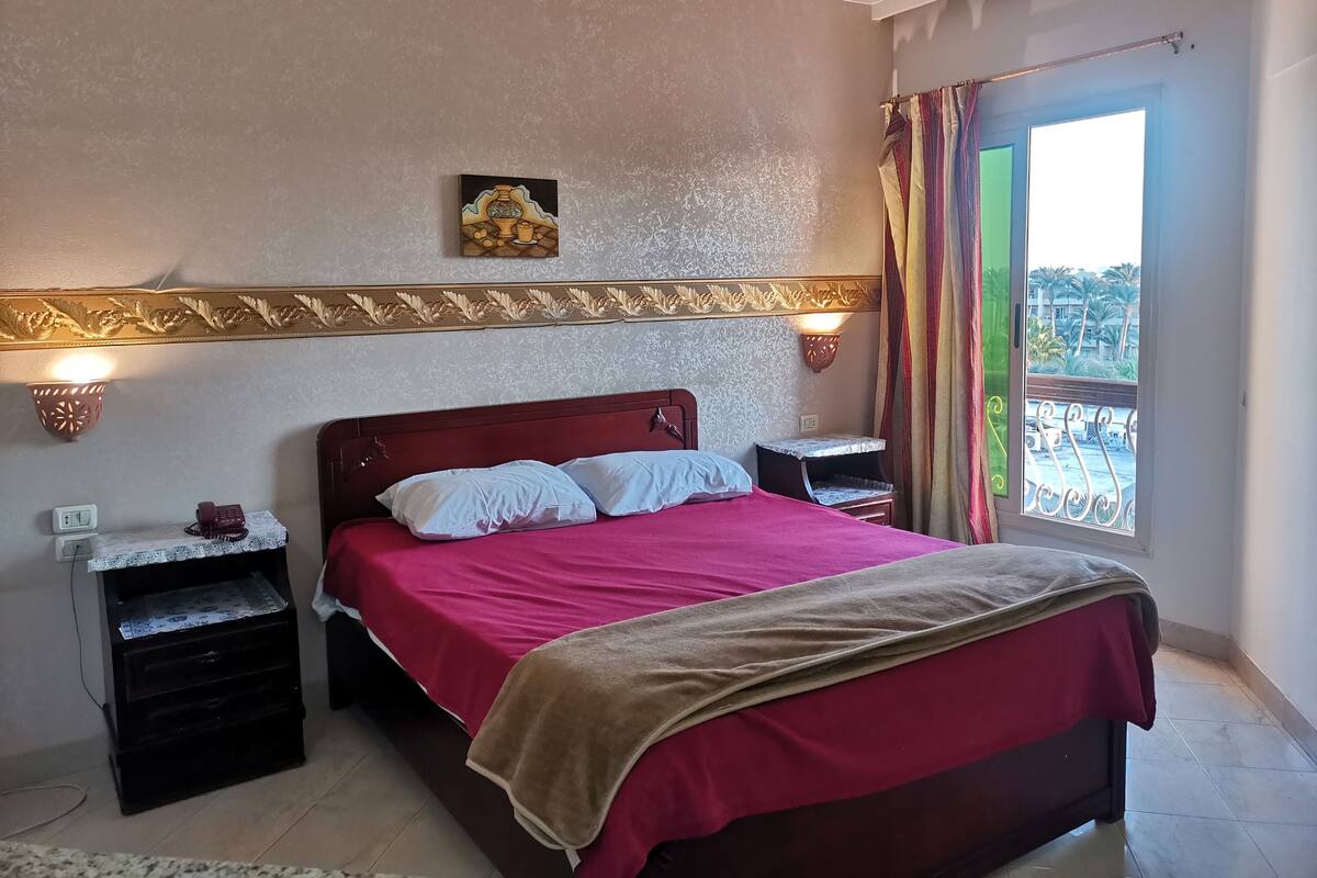 A bedroom with a bed, bedside tables, a wall-mounted lamp, a decorative wall piece, and a view of the outside.
