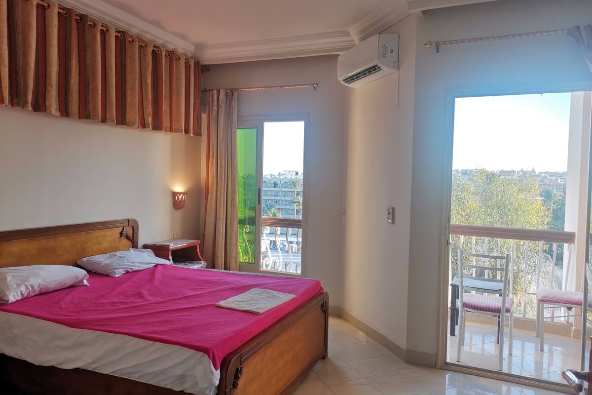 A hotel room with a bed, a nightstand, a wall-mounted air conditioner, a balcony with chairs, and a view of the city.