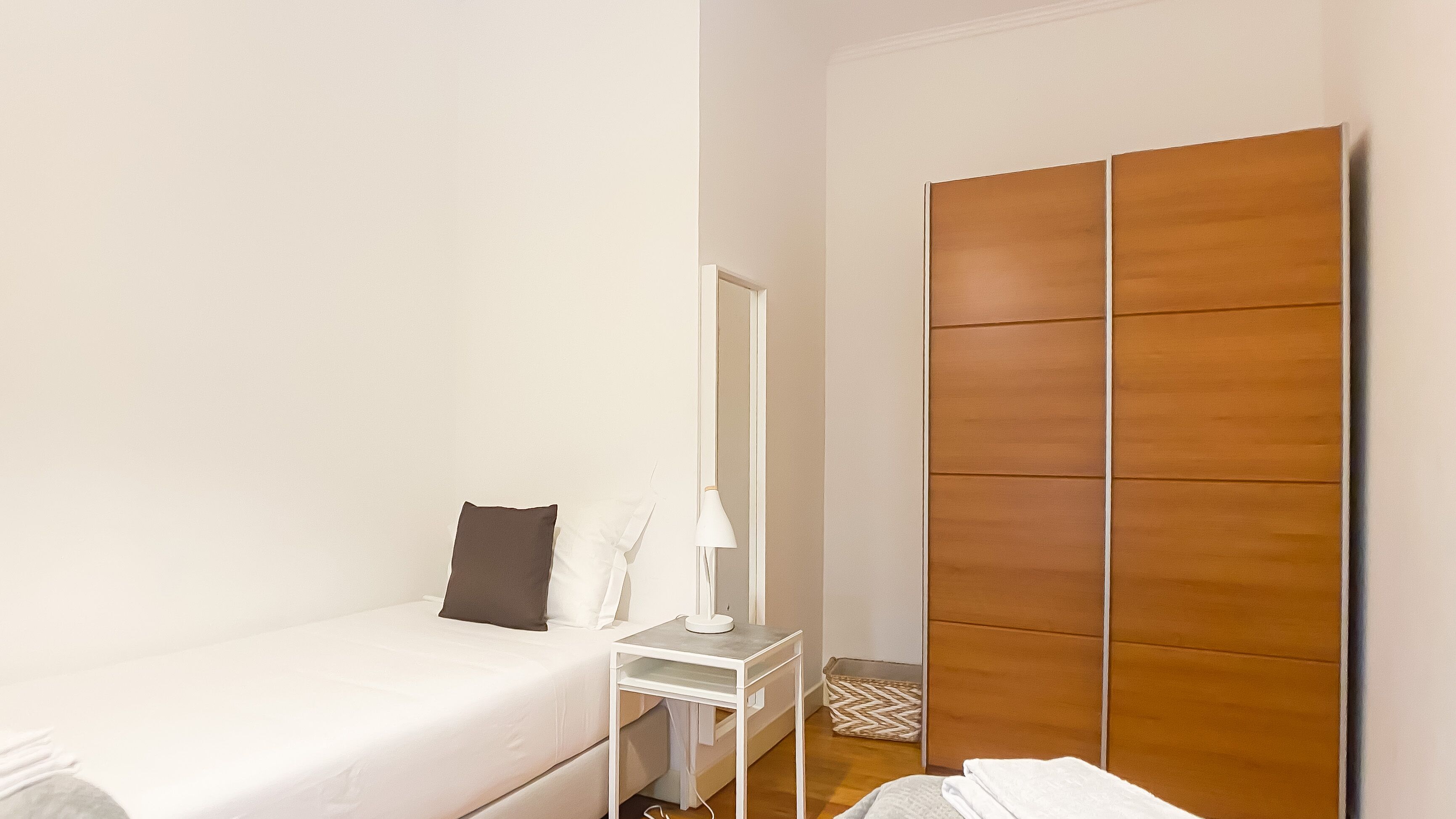 Une petite chambre à coucher, bien rangée, avec un lit simple, une table de chevet avec une lampe, une armoire et un petit banc.