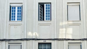 Eine Gebäudefassade mit mehreren Fenstern, davon einige mit Fensterläden, und einer zentralen Tür mit einem Schild darüber.