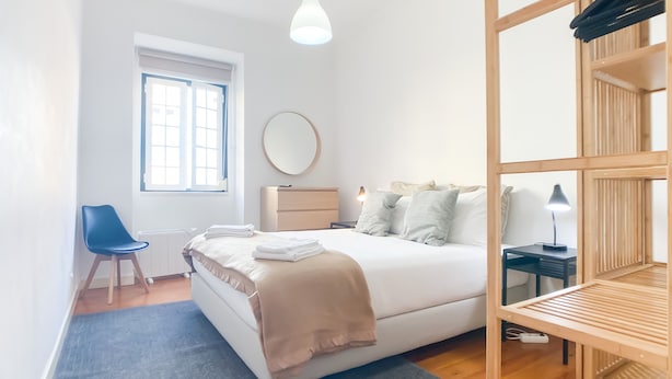 Ein modernes Schlafzimmer mit einem großen Bett, einem Nachttisch mit Lampe, einem Sessel, einer Kommode und einem Fenster mit Jalousien.
