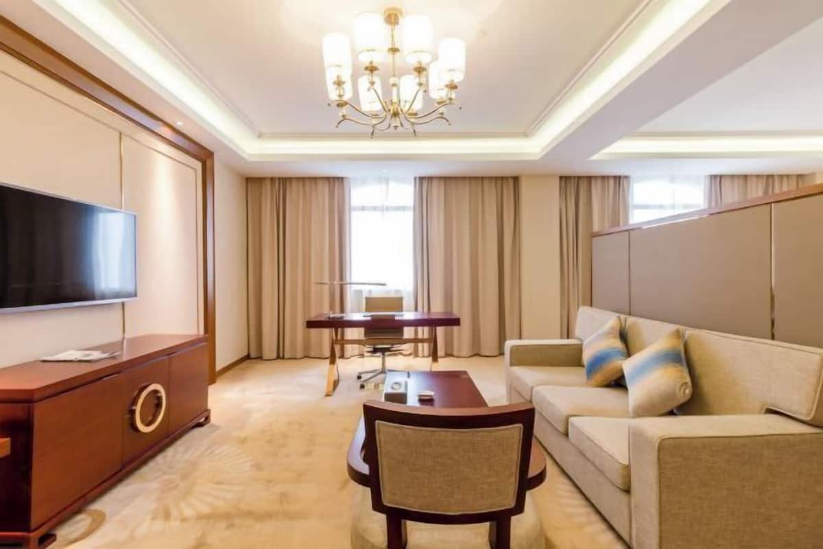 A hotel room with a flat-screen TV, a wooden cabinet, a dining table with chairs, a sofa, and a chandelier.