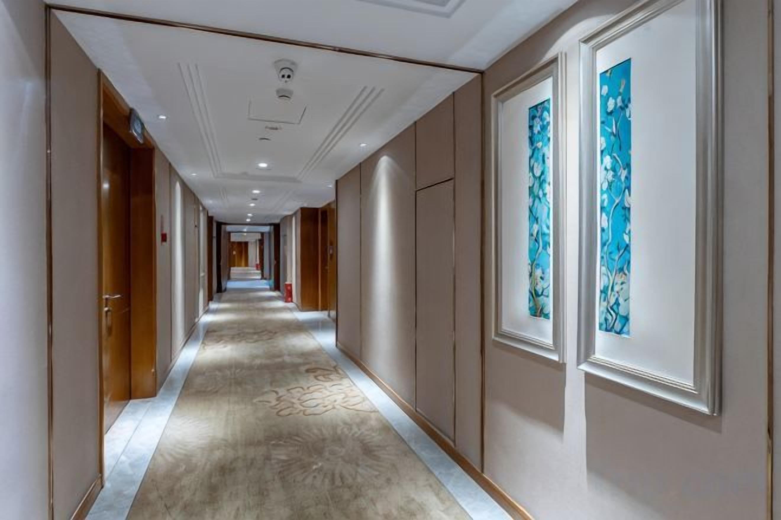 A hotel corridor with doors on both sides, a carpeted floor, and framed artwork on the walls.