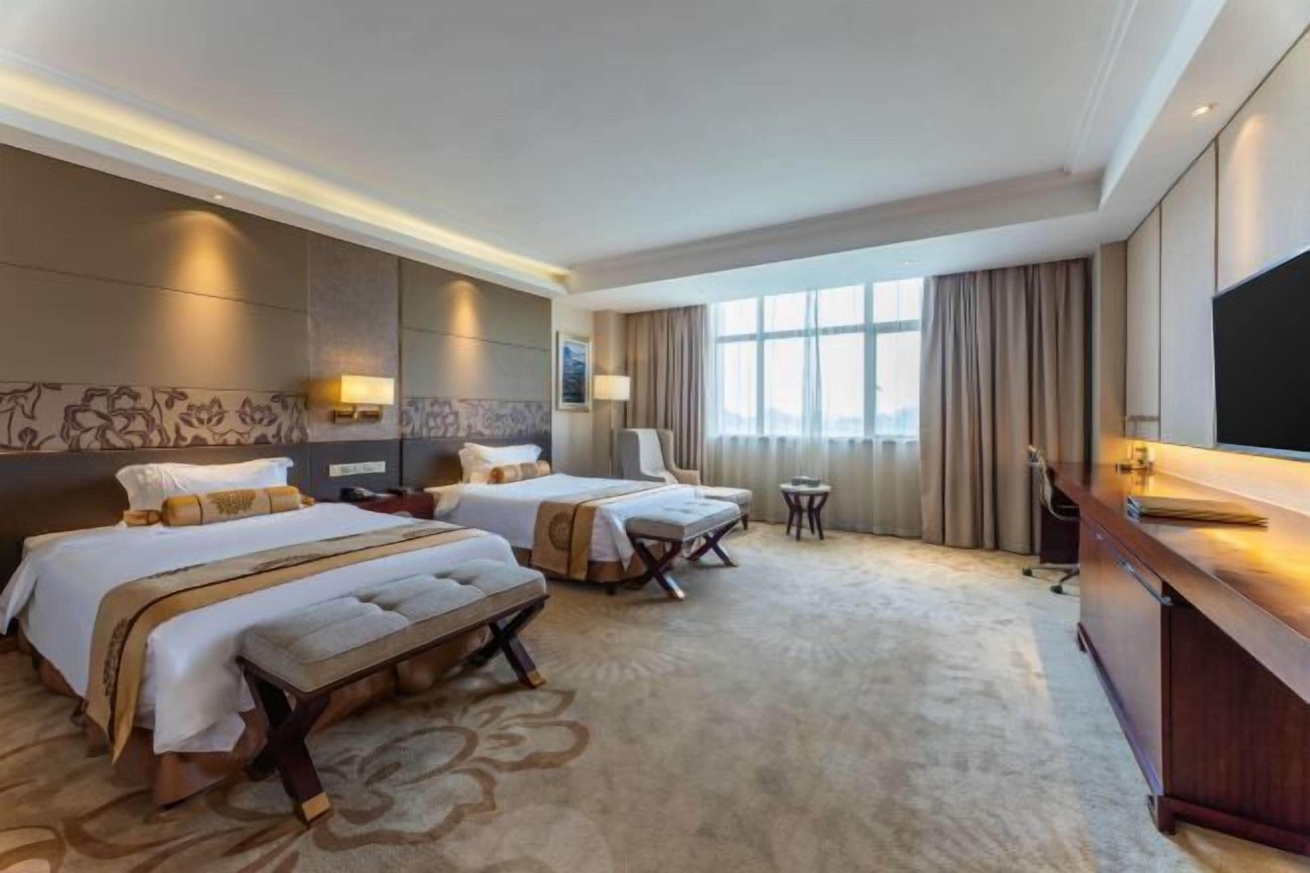 Quarto de hotel com duas camas, uma escrivaninha, uma televisão e uma janela grande.