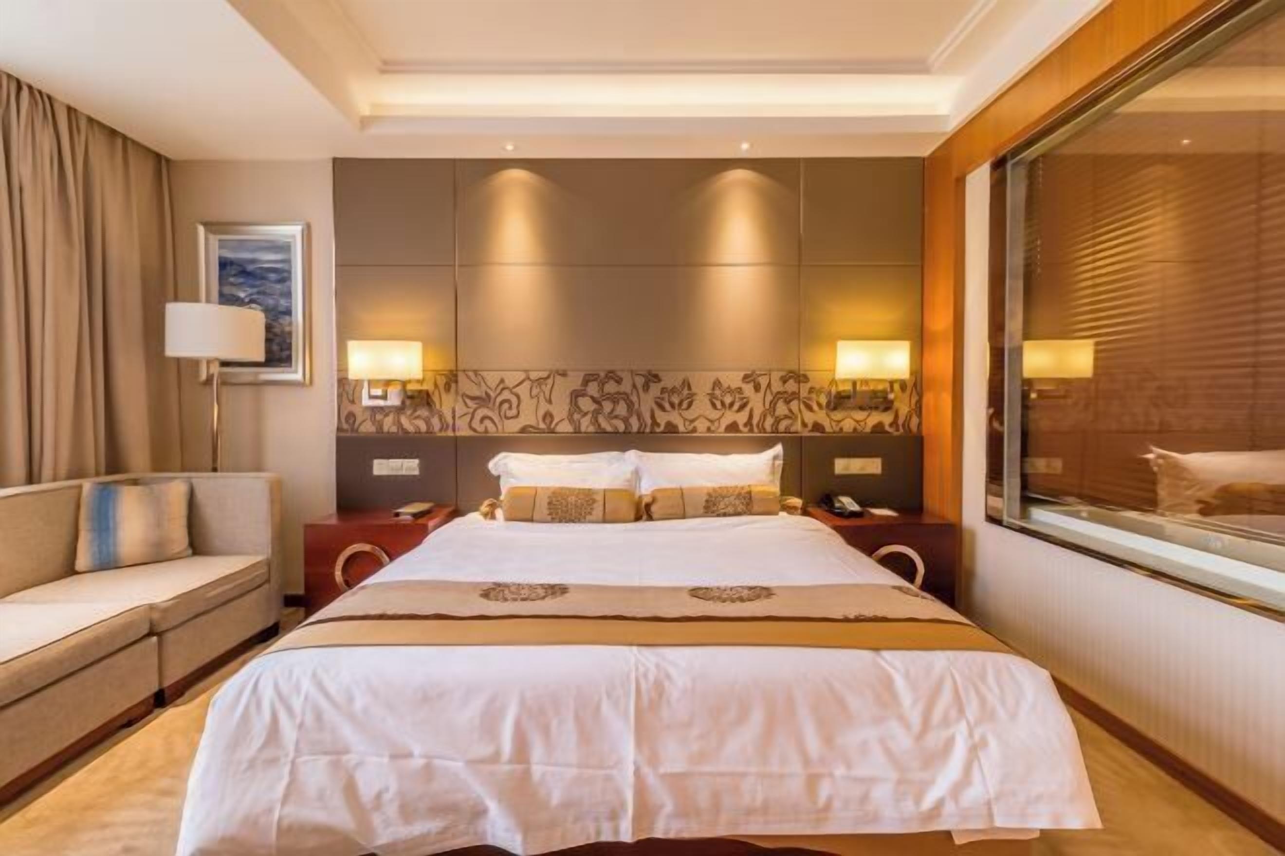 A hotel room with a large bed, a sofa, two bedside tables, and a large window.