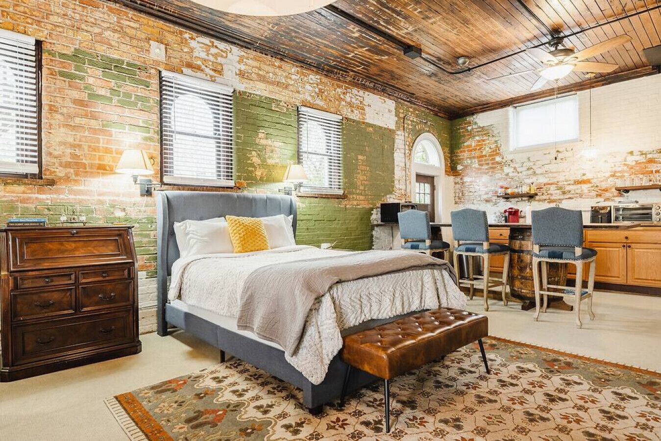 A bedroom with a bed, dresser, and bench, a kitchenette with bar stools, and a brick wall.