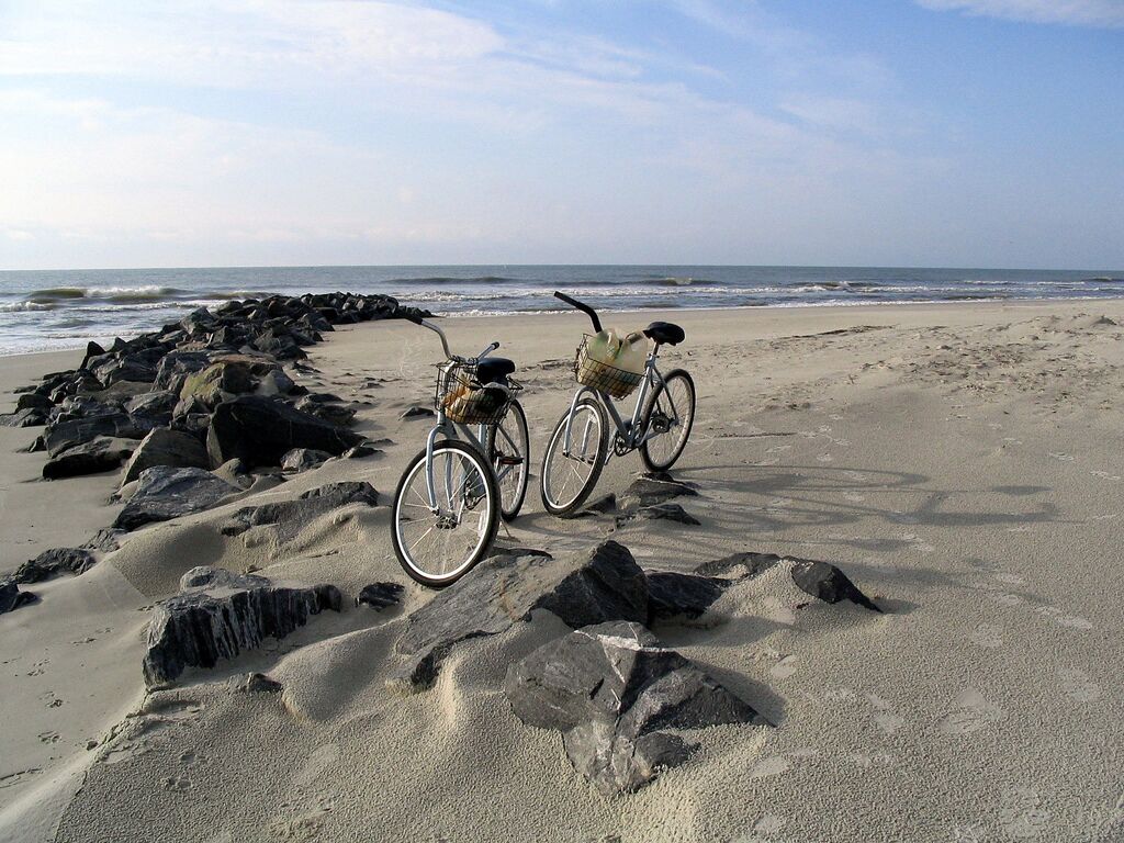 Duas bicicletas estacionadas numa praia de areia com um litoral rochoso e o oceano ao fundo.