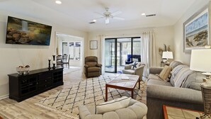 A spacious living room with a ceiling fan, a large TV mounted on the wall, a black entertainment unit, a grey sofa, a patterned rug, and a dining area with a table and chairs. - SeaPines Cheerful Pet Friendly Home by the Beach (Hilton Head Island)