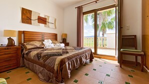 A hotel room with a large bed, wooden headboard, two bedside tables with lamps, a view of palm trees through a sliding glass door, and a balcony.