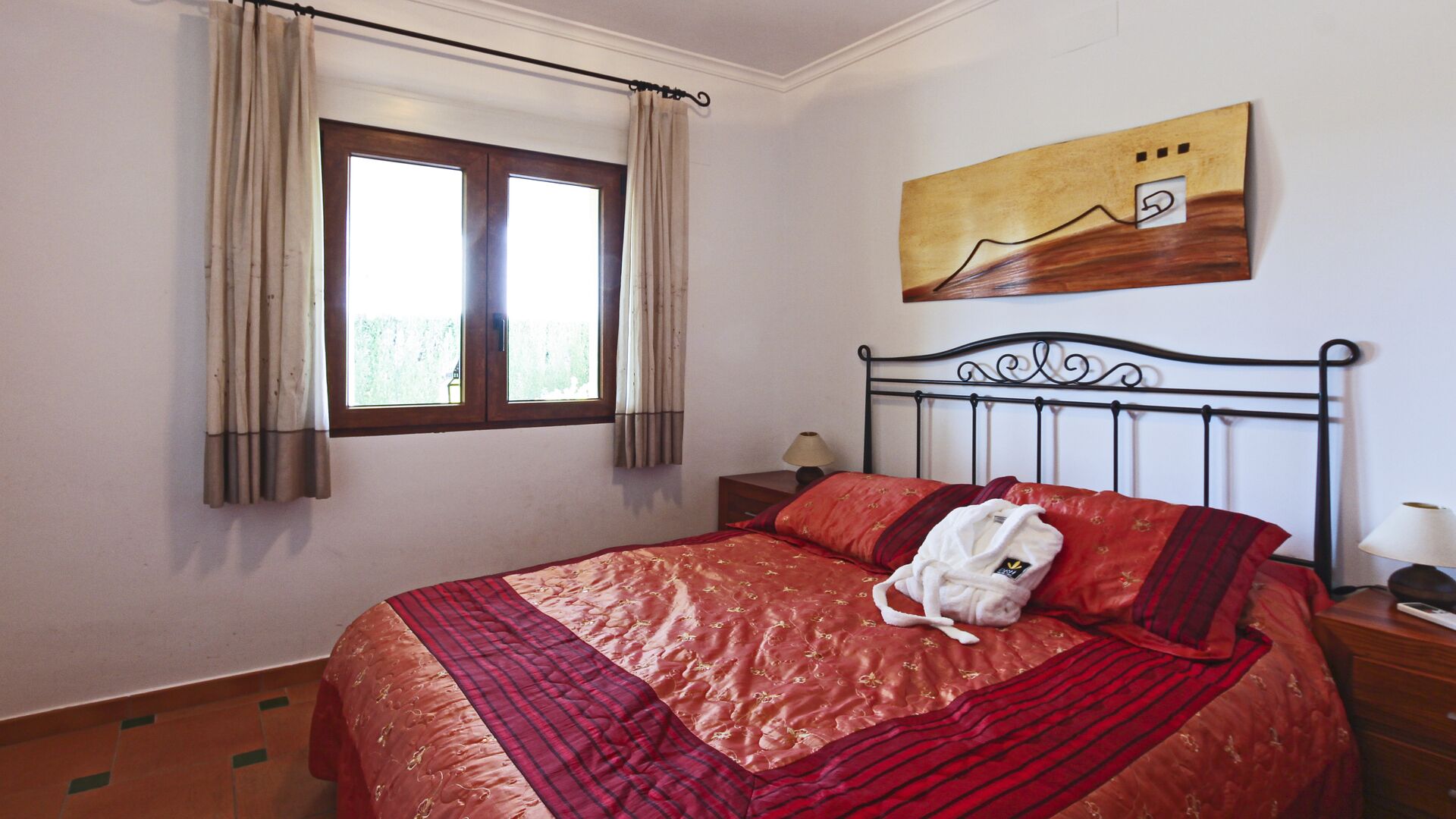 A bedroom with a bed, a window with curtains, a painting on the wall, and a nightstand with a lamp.