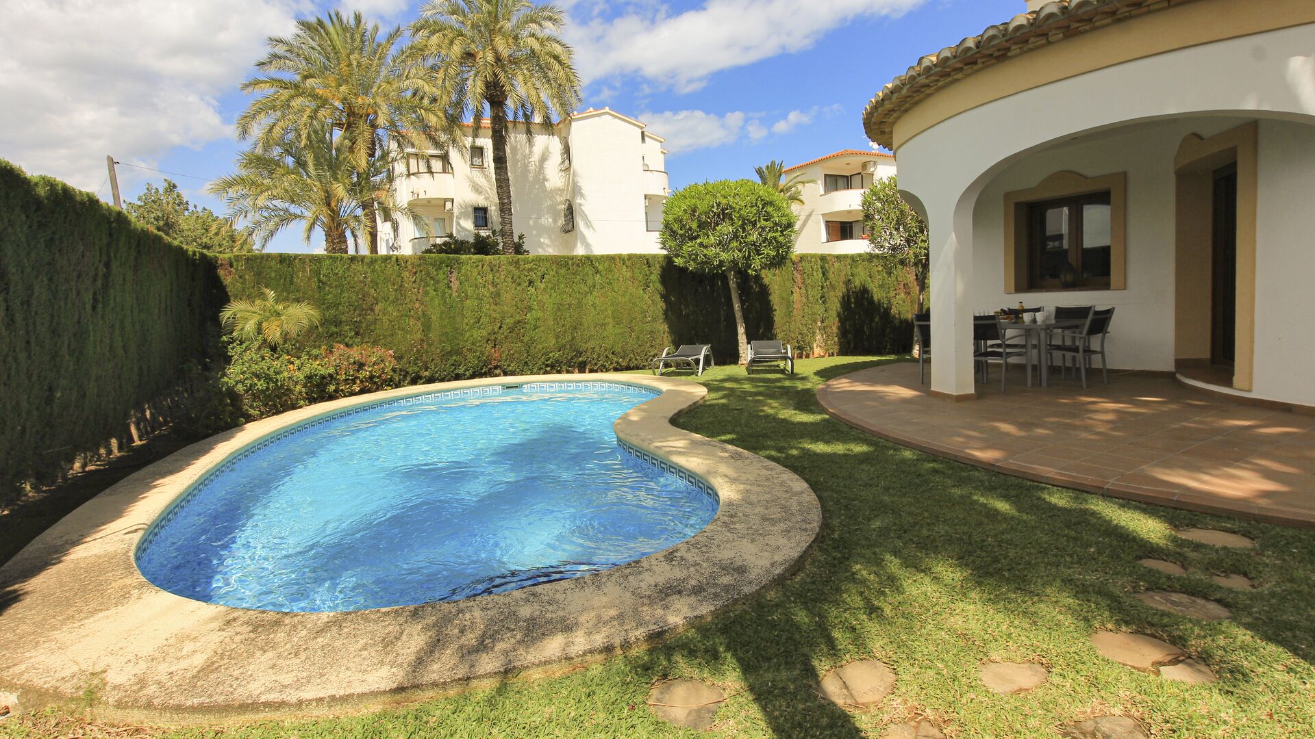A swimming pool with a curved edge, surrounded by a well-maintained lawn and a hedge. There are lounge chairs and a patio area with a table and chairs.